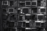 Computer monitors, with the screen turned off, on row of shelves. Opened desktops are next to some of the monitors.